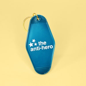 Pu&ograve; includere: Un portachiavi blu a forma di chiave di motel con un anello dorato e il testo "the anti-hero" con tre stelle bianche.