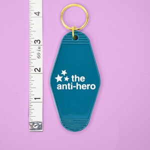 Pu&ograve; includere: Portachiavi da motel blu turchese con un anello per chiavi dorato. Il portachiavi ha tre stelle bianche e il testo "the anti-hero" in bianco.
