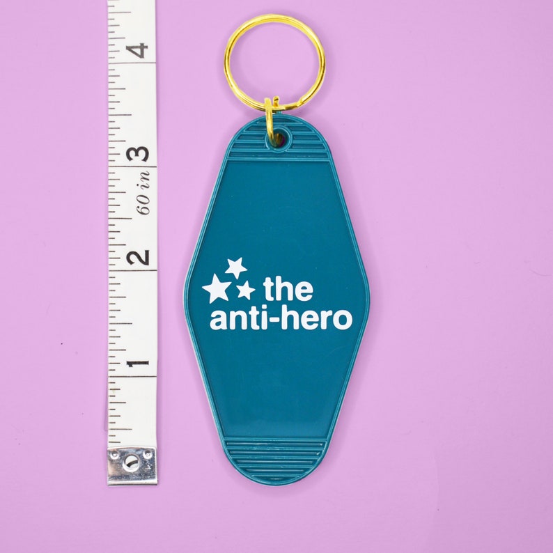 Pu&ograve; includere: Portachiavi da motel blu turchese con un anello per chiavi dorato. Il portachiavi ha tre stelle bianche e il testo "the anti-hero" in bianco.