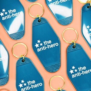 Pu&ograve; includere: Un set di portachiavi a forma di chiave di motel blu turchese con anelli dorati. Ogni portachiavi presenta stelle bianche e il testo "the anti-hero".