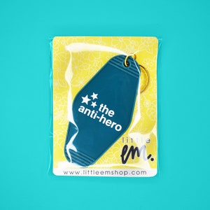 Pu&ograve; includere: Un portachiavi a forma di chiave di motel blu turchese con il testo "the anti-hero" e due stelle. Il portachiavi &egrave; confezionato in un sacchetto di plastica trasparente con sfondo giallo.