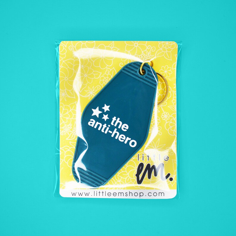 Pu&ograve; includere: Un portachiavi a forma di chiave di motel blu turchese con il testo "the anti-hero" e due stelle. Il portachiavi &egrave; confezionato in un sacchetto di plastica trasparente con sfondo giallo.