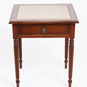 Op de afbeelding: Kleine, antieke houten bijzettafel met een vierkant blad en een enkele lade. De tafel heeft een donkerbruine afwerking, een lichtgekleurde lederen inzet op het tafelblad en decoratief gesneden poten. De lade heeft een ronde messing knop. De tafel is ongeveer 60 cm hoog.
