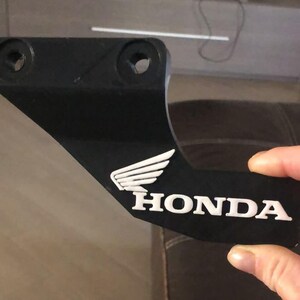 Può includere: Pezzo di moto nero con un logo Honda bianco e un design ad ala bianca.