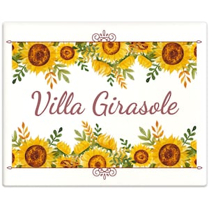 Könnte beinhalten: Ein weißes Schild mit braunem Rand und einem floralen Design aus gelben Sonnenblumen und grünen Blättern. Der Text "Villa Girasole" ist in brauner Kursivschrift geschrieben.