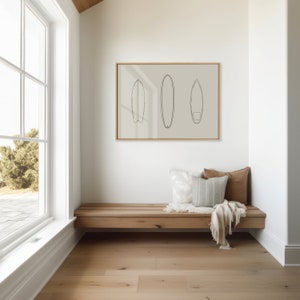 May include: A framed print of three line drawings of surfboards in black on a light beige background. The print is hanging on a white wall above a wooden bench with two pillows and a throw blanket.