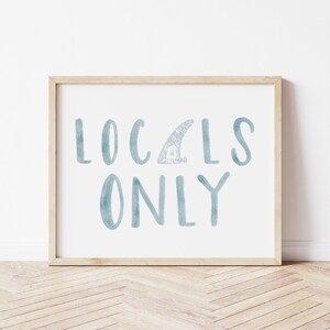 May include: A framed print with the text "LOCALS ONLY" in blue watercolor lettering. A small surfboard fin is drawn in the center of the text.