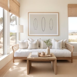 May include: A white couch with three pillows and a wooden frame sits in front of a window with a view of trees. A wooden coffee table with a tray and a mug sits in front of the couch. A framed print of three surfboards hangs on the wall above the couch.