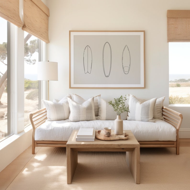 May include: A white couch with three pillows and a wooden frame sits in front of a window with a view of trees. A wooden coffee table with a tray and a mug sits in front of the couch. A framed print of three surfboards hangs on the wall above the couch.