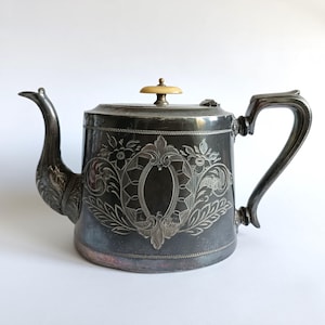Puede incluir: Una tetera de plata con un diseño grabado ornamentado y una perilla de color crema en la tapa.