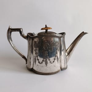Peut inclure: Une théière en argent avec des détails floraux et de guirlandes ornés. La théière présente un bec verseur incurvé, une poignée carrée et un couvercle avec un bouton en bois. La théière est posée sur un fond blanc.