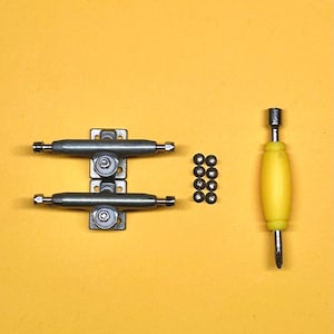 May include: A set of silver-colored skateboard trucks, eight small black bearings, and a yellow screwdriver on a yellow background. The trucks are mounted on a silver plate. The screwdriver has a silver tip.