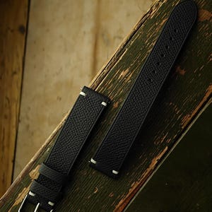 Könnte beinhalten: Zweiteiliges schwarzes Uhrenarmband mit weißen Nähten. Das Lederarmband hat eine strukturierte Oberfläche und eine silberne Schnalle. Das Armband wird auf einer verwitterten, grünen Holzoberfläche präsentiert.