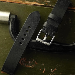 Może przedstawiać: Dwa czarne skórzane paski do zegarków ze srebrnymi sprzączkami. Jeden pasek ma wiele otworów do regulacji. Paski leżą na zielonej, teksturowanej powierzchni. Paski do zegarków to klasyczny dodatek.