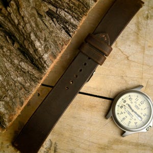 Op de afbeelding: Een bruine leren horlogeband met een zilveren gesp en stiksels. De band ligt naast een zilveren horloge met een witte wijzerplaat en zwarte cijfers. De achtergrond is een lichtbruin houten oppervlak.