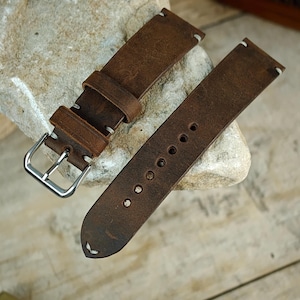 May include: Two-piece brown leather watch strap with white stitching. The strap features a silver buckle and multiple adjustment holes. The leather has a distressed appearance, suggesting a vintage or rustic style. The strap is designed to attach to a watch case.