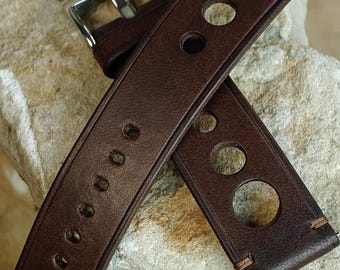 Könnte beinhalten: Ein braunes Leder-Uhrenarmband mit einer silbernen Schnalle. Das Armband hat kreisförmige Aussparungen und Nahtdetails. Das Uhrenarmband wird auf einer strukturierten, hellbraunen Oberfläche gezeigt.