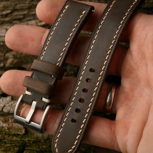 May include: A pair of brown leather watch straps with white stitching. The straps have a stainless steel buckle and are displayed against a textured wooden background. The watch straps are new.