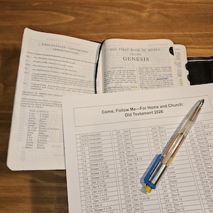 May include: An open Bible with the text "Genesis" visible, alongside a printed study guide titled "Come, Follow Me - For Home and Church: Old Testament 2026." A pen rests on the guide. The scene is set on a wooden surface.