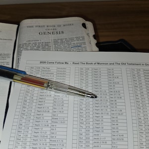 May include: An open book with the title "Genesis" and a reading schedule titled "2026 Come Follow Me." A clear pen with a blue cap rests on the pages.