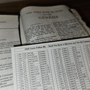 Könnte beinhalten: Ein offenes Buch mit dem Text "THE FIRST BOOK OF MOSES CALLED GENESIS" und einem Leseplan mit dem Titel "2026 Come Follow Me - Read The Book of Mormon and The Old Testament in One Year".