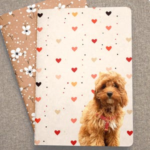 May include: Two notebooks. One has a brown cover with white floral patterns. The other has a cream cover with red, pink, and black hearts and a brown dog. The dog has a red collar.