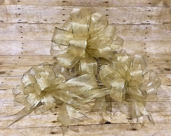 Puede incluir: Tres lazos decorativos dorados con ribete plateado. Los lazos están hechos de una tela transparente y brillante y están dispuestos sobre una superficie de madera. Los lazos son de diferentes tamaños y tienen múltiples bucles y colas.