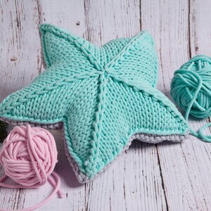 May include: A teal blue knitted star-shaped pillow with a gray border. Two balls of yarn, one teal blue and one pink, are next to the pillow.