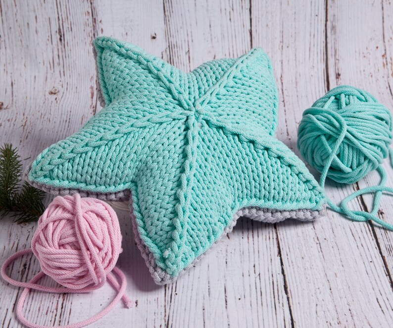 May include: A teal blue knitted star-shaped pillow with a gray border. Two balls of yarn, one teal blue and one pink, are next to the pillow.