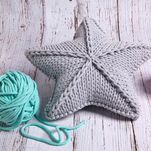 May include: A gray knitted star-shaped pillow with a light blue ball of yarn and a pink ball of yarn on a white wooden background.
