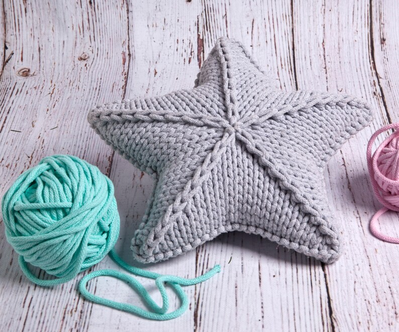 May include: A gray knitted star-shaped pillow with a light blue ball of yarn and a pink ball of yarn on a white wooden background.