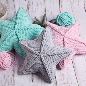 May include: Three knitted star-shaped pillows in pink, gray, and light blue. The pillows are made with chunky yarn and have a textured surface. There are balls of yarn in pink and light blue in the image.