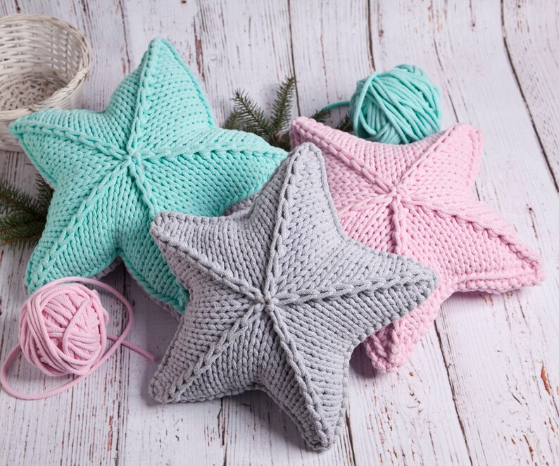 May include: Three knitted star-shaped pillows in pink, gray, and light blue. The pillows are made with chunky yarn and have a textured surface. There are balls of yarn in pink and light blue in the image.