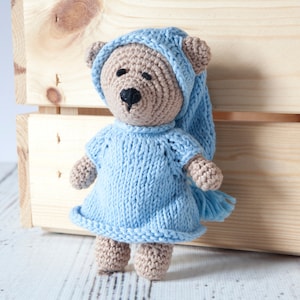 Op de afbeelding: Een gehaakte teddybeer die een blauwe gebreide nachtjapon en een blauwe gehaakte slaapmuts draagt. De beer staat op een houten oppervlak.