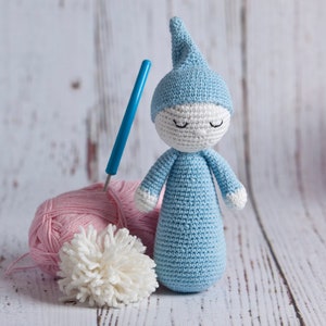 Puede incluir: Una muñeca de crochet azul con una cara blanca y los ojos cerrados. La muñeca lleva un gorro de crochet azul. La muñeca está sentada sobre una superficie de madera blanca con hilo rosa y un pompón blanco.