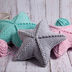 May include: Three knitted star-shaped pillows in different colors: light blue, gray, and pink. The pillows are made with thick yarn and have a textured surface.