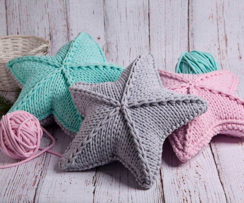 May include: Three knitted star-shaped pillows in different colors: light blue, gray, and pink. The pillows are made with thick yarn and have a textured surface.