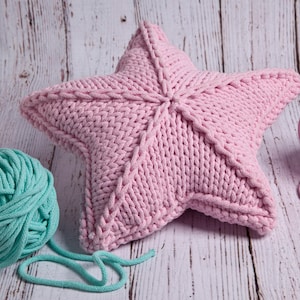 May include: A pink knitted star-shaped pillow on a white wooden surface. There is a ball of teal yarn and a length of teal yarn next to the pillow.