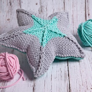 May include: A gray and teal knitted star-shaped pillow with two balls of yarn, one pink and one teal, on a wooden surface.