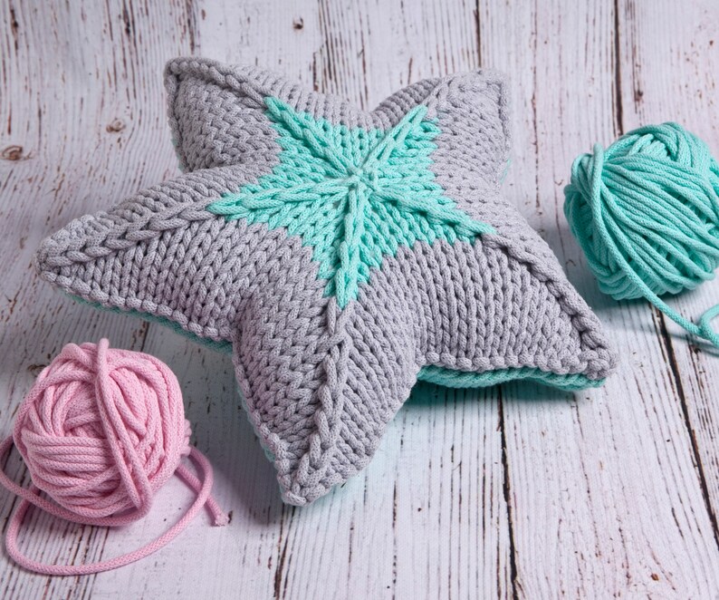 May include: A gray and teal knitted star-shaped pillow with two balls of yarn, one pink and one teal, on a wooden surface.