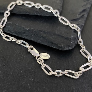 May include: A silver chain bracelet with an oval link design. The bracelet has a lobster clasp closure and a small tag with text. The bracelet is displayed on a dark, textured surface.