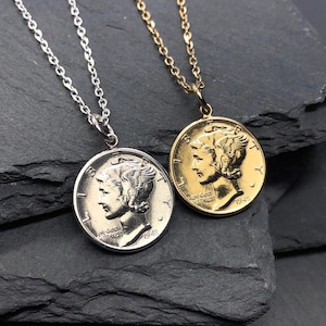 May include: Two coin pendant necklaces, one silver-toned and one gold-toned, each featuring a profile of a person. The silver necklace has a silver chain, and the gold necklace has a gold chain. The coins are set against a dark grey, textured background.