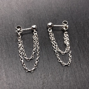 May include: A pair of silver chain earrings. Each earring features a small ball stud and two delicate chains that hang down. The earrings are displayed on a dark gray surface.