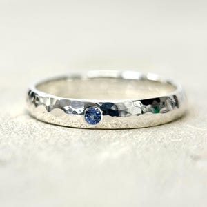May include: A silver ring with a hammered texture and a small, round, blue gemstone. The ring is set against a neutral background, highlighting its simple elegance and craftsmanship. The gemstone adds a subtle pop of color.