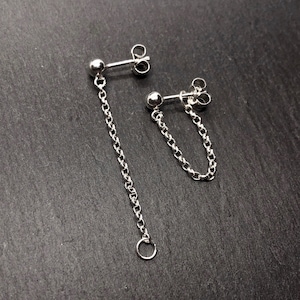 Peut inclure: Une paire de boucles d'oreilles en chaîne argentées. L'une comporte une longue chaîne avec un petit anneau, l'autre une chaîne plus courte. Les deux ont un petit clou à boule en haut. Sur fond gris foncé.