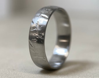 May include: A silver-colored ring with a textured surface. The ring has a band width of approximately 0.25 inches. The surface of the ring has a unique, organic pattern, adding visual interest to the piece. The ring is likely made of metal.