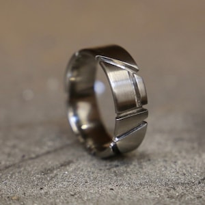 May include: A silver-colored ring with a geometric design. The ring features a series of angled and straight cuts, creating a modern, architectural look. The metal has a brushed finish, giving it a matte appearance. The ring is likely made of stainless steel or a similar metal.
