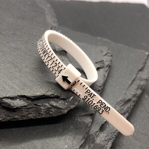May include: A white plastic ring sizer with black markings. The sizer has letters and numbers, and an arrow indicating the size. The text "PAT. PEND. 9101693" is printed on the sizer.