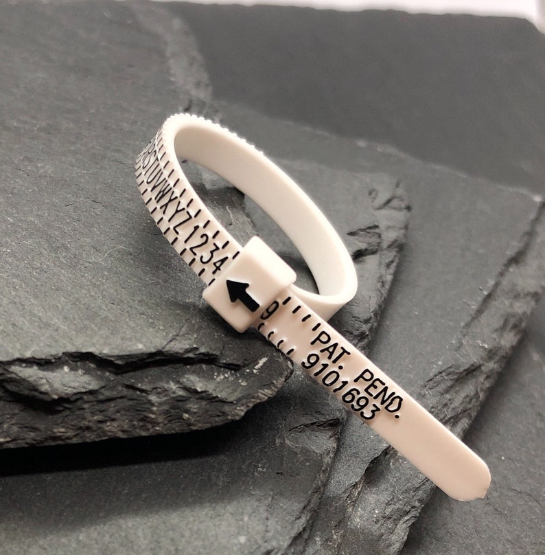 May include: A white plastic ring sizer with black markings. The sizer has letters and numbers, and an arrow indicating the size. The text "PAT. PEND. 9101693" is printed on the sizer.