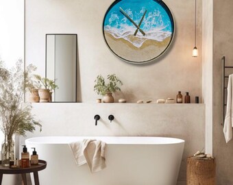 May include: A round wall clock with a beach scene design. The clock face features a blue ocean, white waves, and tan sand. The clock has black hands and a silver frame. The clock is mounted on a beige wall in a bathroom setting.